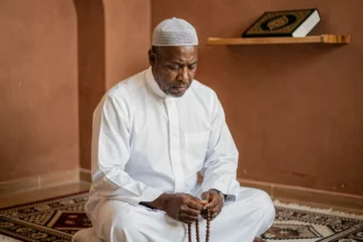 Homme en tenue de prière assis sur un tapis de prière dans une salle de mosquée à Rennes, tenant un chapelet en contemplation silencieuse
